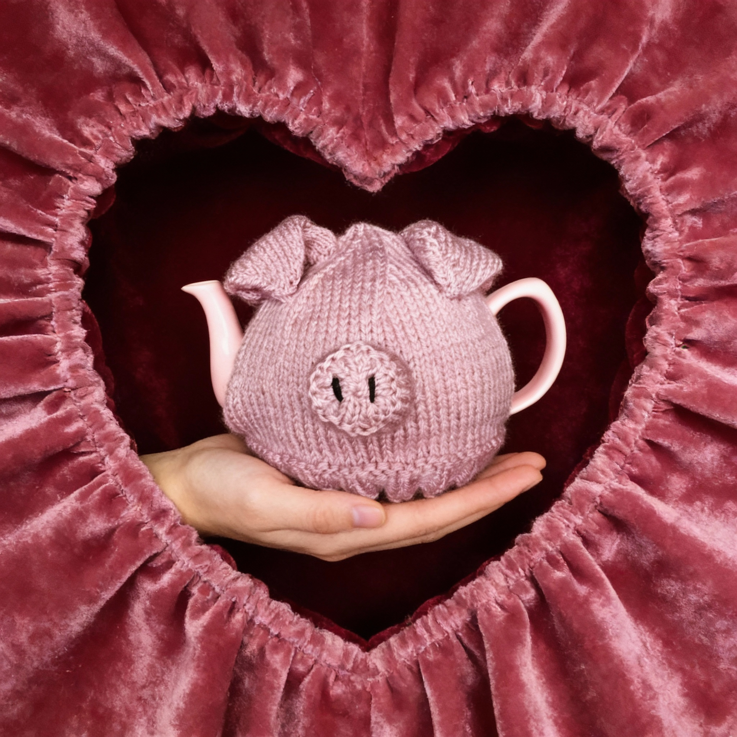 Pink knitted pig-shaped teapot held by hands against a heart-shaped pink velvet background